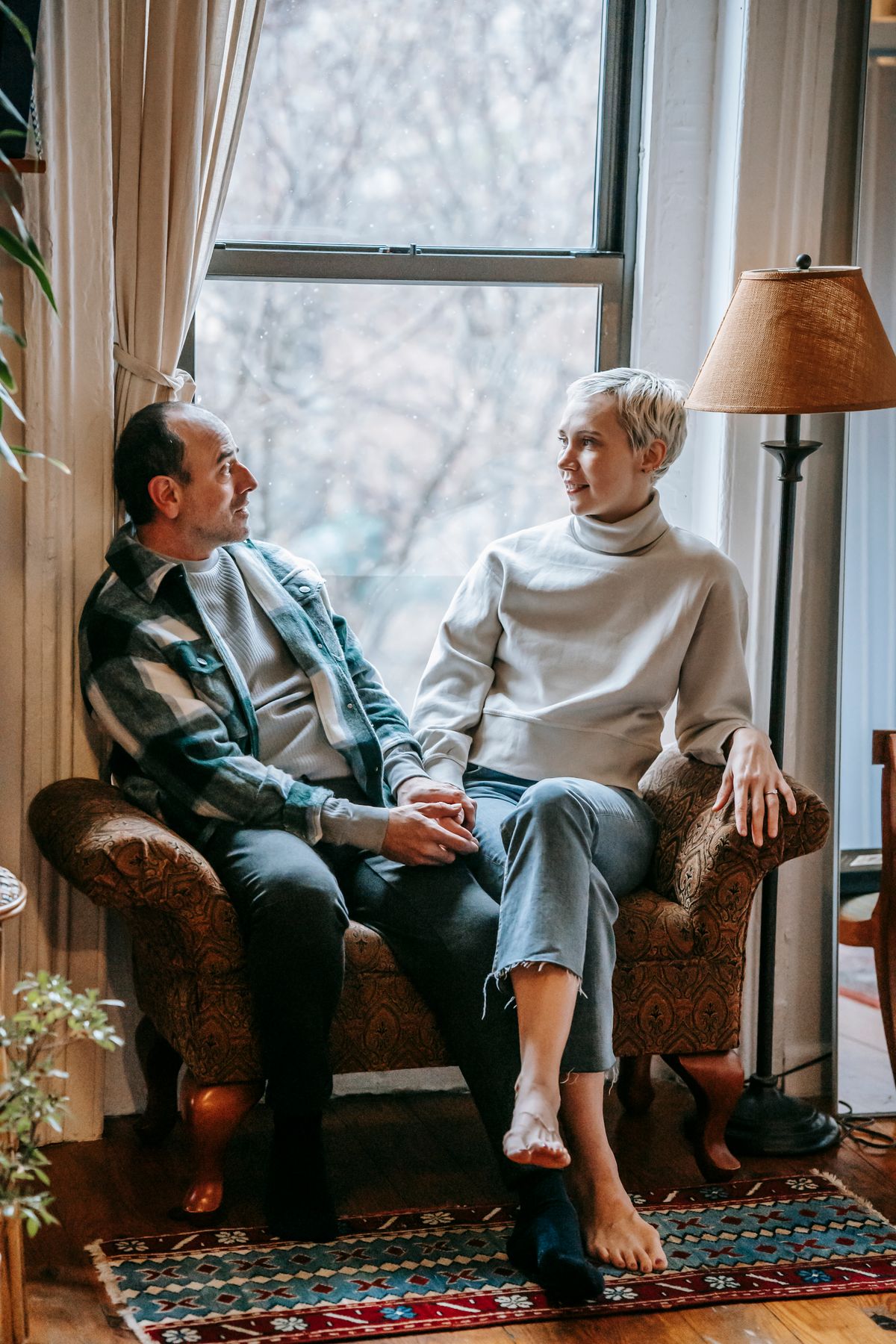 A husband and wife sitting close together by a window, holding hands and talking